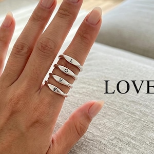 Könnte beinhalten: Vier silberfarbene Ringe, die an einem Finger gestapelt sind und jeweils einen schwarzen Buchstaben tragen, sodass das Wort LOVE entsteht. Die Ringe haben eine glatte, polierte Oberfläche und ein schlichtes Design. Ein goldenes Kettenarmband mit Sternanhänger ist zu sehen.