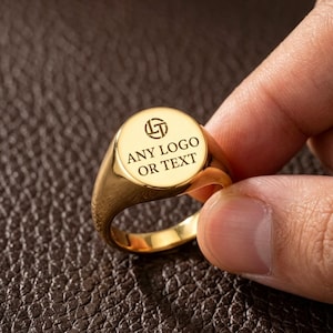 Peut inclure: Une bague chevalière dorée avec une face plate circulaire présentant une icône de logo circulaire et le texte gravé ANY LOGO OR TEXT. La bague possède une finition métallique polie et un design d'anneau lisse et arrondi, offrant un aspect épuré.