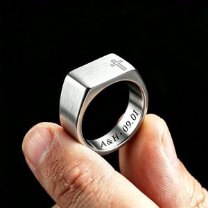 May include: A silver-toned metal signet ring featuring a brushed finish on the flat top surface with an engraved cross symbol. The inner band of the ring is engraved with the text A&H • 09.01 in a clear, dark, sans-serif font style.