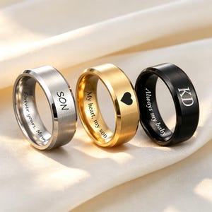 May include: Three metal rings in silver, gold, and black tones. The silver ring is engraved with 'SON' and 'Forever yours, Mom'. The gold ring features a heart icon and 'My heart, my son'. The black ring displays 'KD' and 'Always my baby'.