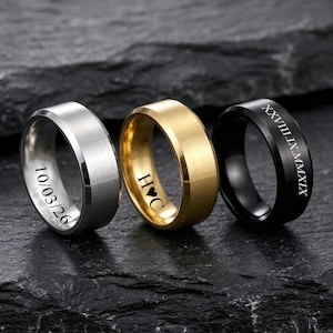 May include: Three metal rings with a brushed finish in silver, gold, and black. The silver ring is engraved with 10/03/26, the gold ring features H heart C, and the black ring displays the Roman numeral date XXVIII.IX.MMXIX on the outer band.