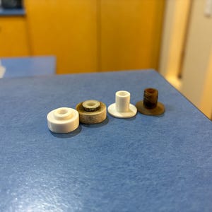 Könnte beinhalten: Vier kleine kreisförmige Bauteile in einer Reihe angeordnet. Zwei sind aus massivem weißem Kunststoff mit einem erhöhten Mittelzylinder, eines ist eine verwitterte beige Scheibe mit weißem Kern, eines ist eine dunkelbraune Scheibe mit weißem Einsatz.
