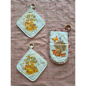 May include: Two diamond-shaped quilted pot holders featuring a yellow and orange graphic of a pepper mill and flowers, and one rectangular oven mitt with a graphic of wheat stalks, a butterfly, and the text WHEAT in orange lettering.