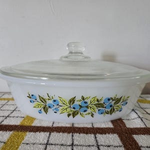 May include: A white oval casserole dish made of opaque glass featuring a decorative pattern of blue berries and green leaves around the exterior. The dish includes a clear, transparent glass lid with a central knob handle on the top.