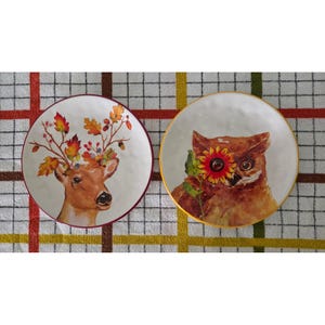 May include: Two round white plates featuring watercolor-style illustrations. The left plate displays a deer with antlers made of autumn leaves and acorns. The right plate shows a brown owl holding a bright yellow sunflower over one of its eyes.