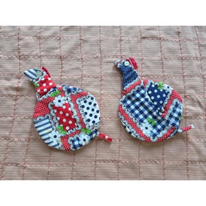 May include: Two round fabric pot holders featuring a patchwork design of red, white, and blue patterns including polka dots, gingham checks, and floral prints. Each piece has a small neck-like extension with a white button and a small fabric loop.