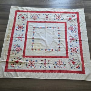 May include: A square off-white tablecloth featuring a red polka-dot border and a central square frame. The fabric is decorated with embroidered motifs of figures in traditional attire, hearts, and floral patterns in red, blue, and green thread.