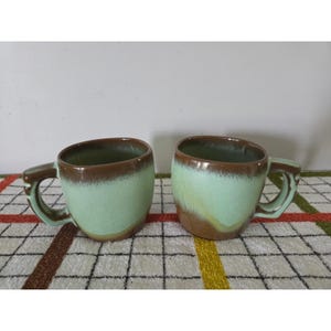 May include: Two ceramic mugs featuring a two-tone glaze with a light mint green body and a dark brown rim that drips down the sides. Each mug has a sturdy, angular handle and a smooth, rounded shape, showcasing a reactive glaze finish.
