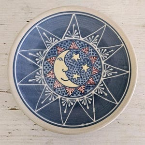 May include: A round ceramic plate with a celestial design. The plate features a dark blue background with a white starburst pattern. The center has a smiling moon and stars in cream, red, and blue.