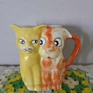 May include: A vintage ceramic pitcher featuring a cat and dog design. The cat is yellow, and the dog is orange and white. The pitcher has a handle and is sitting on a crocheted doily. The pitcher is a unique decorative item.