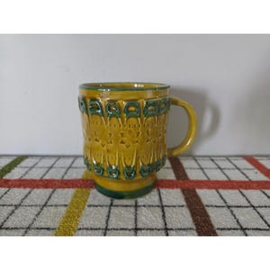 May include: A ceramic mug featuring a mustard yellow glaze with dark green accents. The exterior surface is decorated with a repeating embossed geometric pattern, including stylized floral motifs and vertical arches around the body.