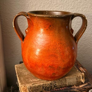 May include: A round, two-handled ceramic vase featuring a vibrant orange and brown glazed finish. The vessel has a wide opening and a smooth, glossy texture with visible signs of wear along the upper rim and the edges of the two curved handles.