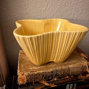 May include: A pale yellow ceramic bowl with a distinct clover-like shape and vertical ribbed detailing along the exterior walls. The bowl features a wide, open top and a smooth, glossy finish that reflects light across its curved, fluted surface.