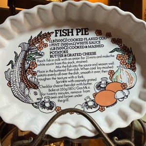 Könnte beinhalten: Eine ovale, weiße Keramik-Auflaufform mit gewelltem Rand, verziert mit einer schwarz-orangen Illustration von Fisch, Pilzen und Zwiebeln. In der Mitte befindet sich ein aufgedrucktes Rezept für Fischpastete mit verschiedenen Zutaten.