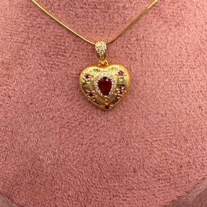 May include: A gold-tone heart-shaped pendant necklace featuring a central pear-cut red stone surrounded by small clear crystals. The heart surface has a textured finish and is accented with additional small red stones and clear crystals.