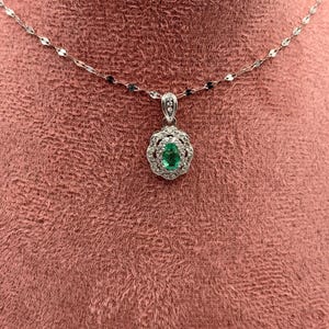 May include: A silver-toned necklace featuring a delicate chain and a pendant with a central oval-cut green gemstone surrounded by a halo of small clear stones, with additional clear stones set along the pendant's bail.