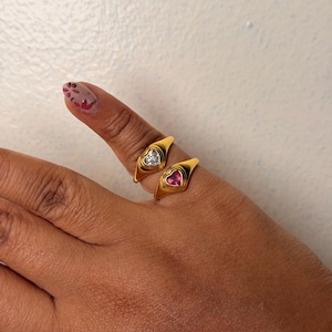 May include: Two gold-toned signet rings featuring heart-shaped stones. The top ring contains a clear, colorless heart-shaped stone, while the bottom ring features a vibrant pink heart-shaped stone. Both rings have a smooth, polished finish.