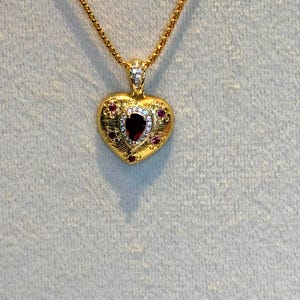 May include: A gold heart-shaped pendant necklace featuring a central pear-shaped dark red gemstone surrounded by a halo of small white stones. The textured gold surface is accented with four small red stones and a single white stone at the bail.