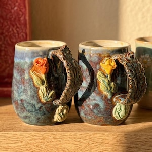 May include: Two ceramic mugs with a blue and brown glaze. Each mug features a sculpted rose and leaf design in orange and yellow, with a decorative handle. The mugs are displayed on a wooden surface.