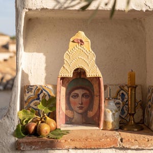 May include: A ceramic shrine with a pointed, textured yellow top and a painted portrait of a person with a teal head covering inside. The shrine is positioned next to a small ceramic pitcher, a brass candle holder with a yellow candle, and fresh figs.