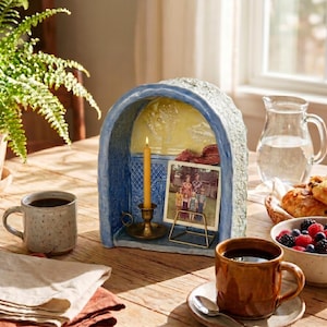 May include: A blue ceramic arched shrine with a textured exterior, featuring a yellow interior back panel, a brass candle holder with a yellow taper candle, and a small gold wire stand displaying a vintage photograph of a family.