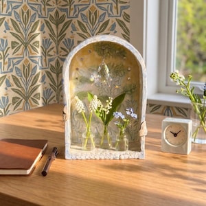 May include: A decorative, arched display shelf with three small glass vases holding white and blue flowers. A crystal prism hangs inside the shelf. A small square clock and a notebook are on the wooden surface.
