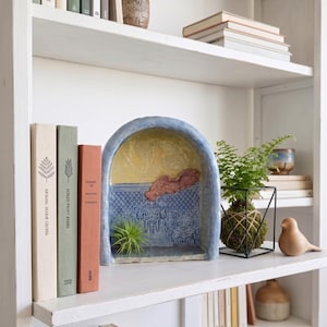 May include: A ceramic arched display niche featuring a yellow upper section and a blue lower section with a textured lattice pattern. A small air plant sits inside the arch, which is positioned next to three books and a green plant in a wire frame.