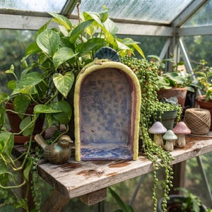 May include: A ceramic arch-shaped display niche with a pale yellow frame and a textured, mottled blue and purple interior. A small lizard figurine rests on the top arch, and a bronze-colored bird figurine sits to the left of the structure.