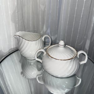 May include: A white porcelain creamer and a lidded sugar bowl set, both featuring a swirling ribbed texture and delicate gold trim along the rims and handles. The sugar bowl includes a matching lid with a small, pointed finial handle.
