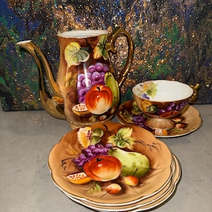 May include: A porcelain tea set featuring a teapot, a teacup, and three saucers, all decorated with hand-painted fruit motifs including grapes, apples, and pears on a tan background, accented with gold trim on the handles and edges.