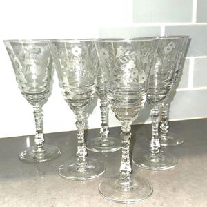 May include: A set of five clear glass goblets featuring etched floral patterns on the bowls. Each goblet has a multi-faceted, decorative stem and a circular base. The glass is transparent, showcasing the intricate botanical engravings on the surface.