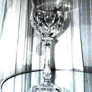 May include: A clear crystal wine glass featuring a deep, intricate cut pattern on the bowl, a multi-faceted stem, and a wide, circular base. The glass reflects light through its geometric carvings, showcasing a polished, transparent finish.