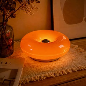 May include: A round, donut-shaped table lamp with a glossy orange finish and a central metallic silver accent. The lamp emits a warm, glowing light from its smooth, curved surface, creating a soft illumination across the immediate area.
