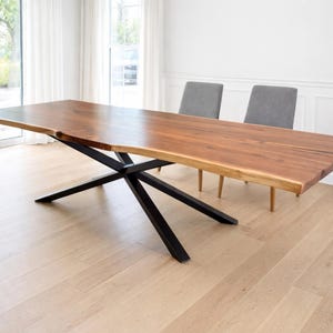 May include: A rectangular dining table featuring a natural live edge wood slab top with a rich brown grain pattern, supported by a black metal base with intersecting diagonal legs, accompanied by two grey upholstered chairs with wooden legs.