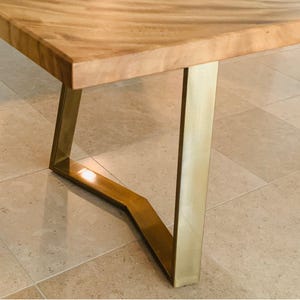 Op de afbeelding: Een tafel met een lichtbruin houten blad met een zichtbaar nerfpatroon en een geometrisch, hoekig metalen onderstel met een geborstelde gouden afwerking. De structuur bestaat uit een plat rechthoekig oppervlak op een stevig frame.