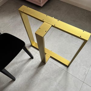 May include: A gold-colored metal table base featuring a rectangular frame design with a central vertical support bar. The top horizontal bar includes multiple pre-drilled circular holes for mounting purposes, providing a stable structure.
