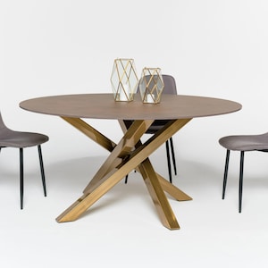May include: A round brown dining table with a gold-toned metal base featuring intersecting legs. Two geometric gold wire candle holders sit on the tabletop. Three grey upholstered chairs with black metal legs are positioned around the table.