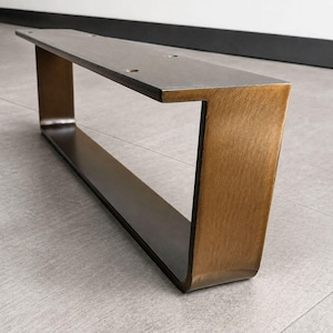 May include: A U-shaped metal furniture leg with a brushed bronze finish. The top horizontal plate features four pre-drilled circular mounting holes for attachment. The metal has a visible grain texture and a warm, metallic brown colour tone.