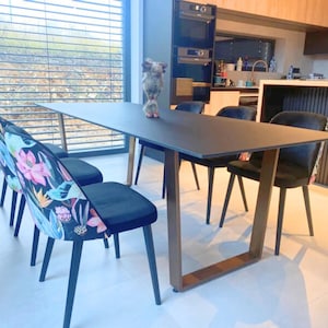 Può includere: Un tavolo da pranzo rettangolare con superficie nera opaca e gambe in metallo color bronzo, accompagnato da quattro sedie con sedute nere e schienali decorati con un vivace motivo floreale di illustrazioni botaniche rosa, blu e verdi.