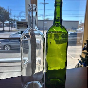 Puede incluir: Dos botellas de vidrio vacías colocadas una al lado de la otra. Una botella es de vidrio transparente, mientras que la otra es de vidrio verde oscuro. Ambas botellas presentan un cuello delgado, una parte superior roscada y un cuerpo cilíndrico.
