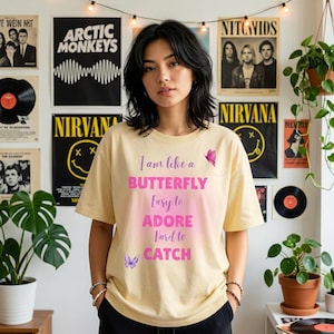 May include: A pale yellow short-sleeve t-shirt featuring the pink text I am like a BUTTERFLY Easy to ADORE Hard to CATCH, accompanied by small purple and pink butterfly illustrations on the front of the garment.