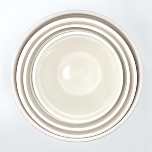 May include: A set of three nested bowls in a smooth, glossy off-white finish. The bowls are arranged in a circular formation, with each bowl fitting inside the next, creating a concentric pattern of rounded rims and deep, reflective interiors.