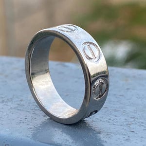 Op de afbeelding: Een gepolijste zilverkleurige metalen ring met een reeks gelijkmatig verdeelde cirkelvormige gravures rond de buitenste band. Elke cirkelvormige gravure bevat een horizontale lijndetail, wat lijkt op een platte schroefkop.