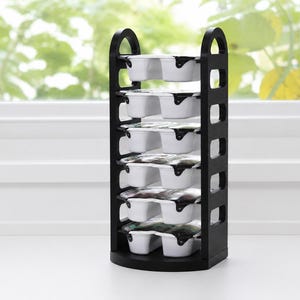 May include: A black vertical storage rack with six stacked levels, each holding a white rectangular container with a dark lid. The rack features arched top handles and side cutouts, providing a structured display for the six individual units.