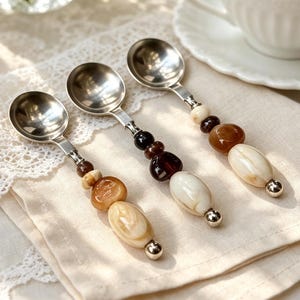 May include: Three silver-toned metal spoons with decorative handles featuring a series of smooth, rounded beads in shades of cream, brown, and dark amber. Each spoon handle ends with a small metallic sphere, showcasing a polished finish.