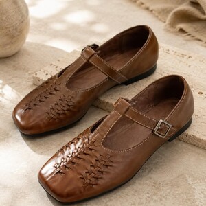 May include: A pair of brown leather T-strap shoes featuring a woven design on the rounded square toe, a thin adjustable ankle strap with a silver-toned metal buckle, white contrast stitching along the edges, and a flat black sole.