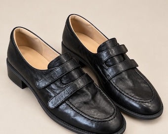 May include: A pair of black leather loafers featuring a textured finish, a rounded toe, and two horizontal straps across the vamp. The shoes have a low block heel, a stitched apron seam, and a light beige interior lining, providing a classic look.