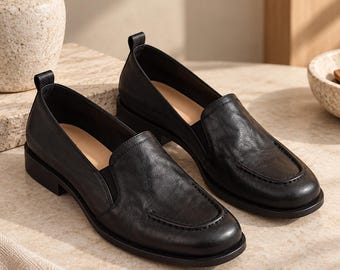 May include: A pair of black leather loafers featuring a smooth, slightly textured finish, a rounded toe, a raised seam detail on the vamp, and a small pull tab at the heel. The interior of the shoes is lined with a light tan material and has a low heel.