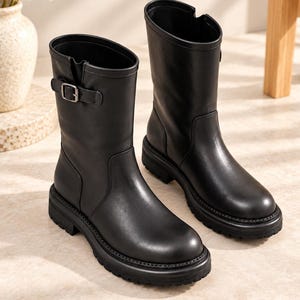 May include: A pair of black mid-calf boots featuring a smooth finish, a rounded toe, and a thick lug sole. Each boot includes a decorative side strap with a silver-toned metal buckle detail near the top collar, providing a structured silhouette.