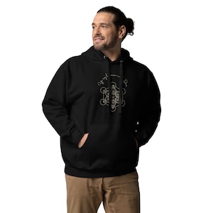 Puede incluir: Una sudadera con capucha negra que presenta un diseño geométrico de color claro en el pecho. El gráfico consiste en un patrón complejo de círculos y líneas que se cruzan, con texto en cursiva arqueado sobre la forma geométrica central.